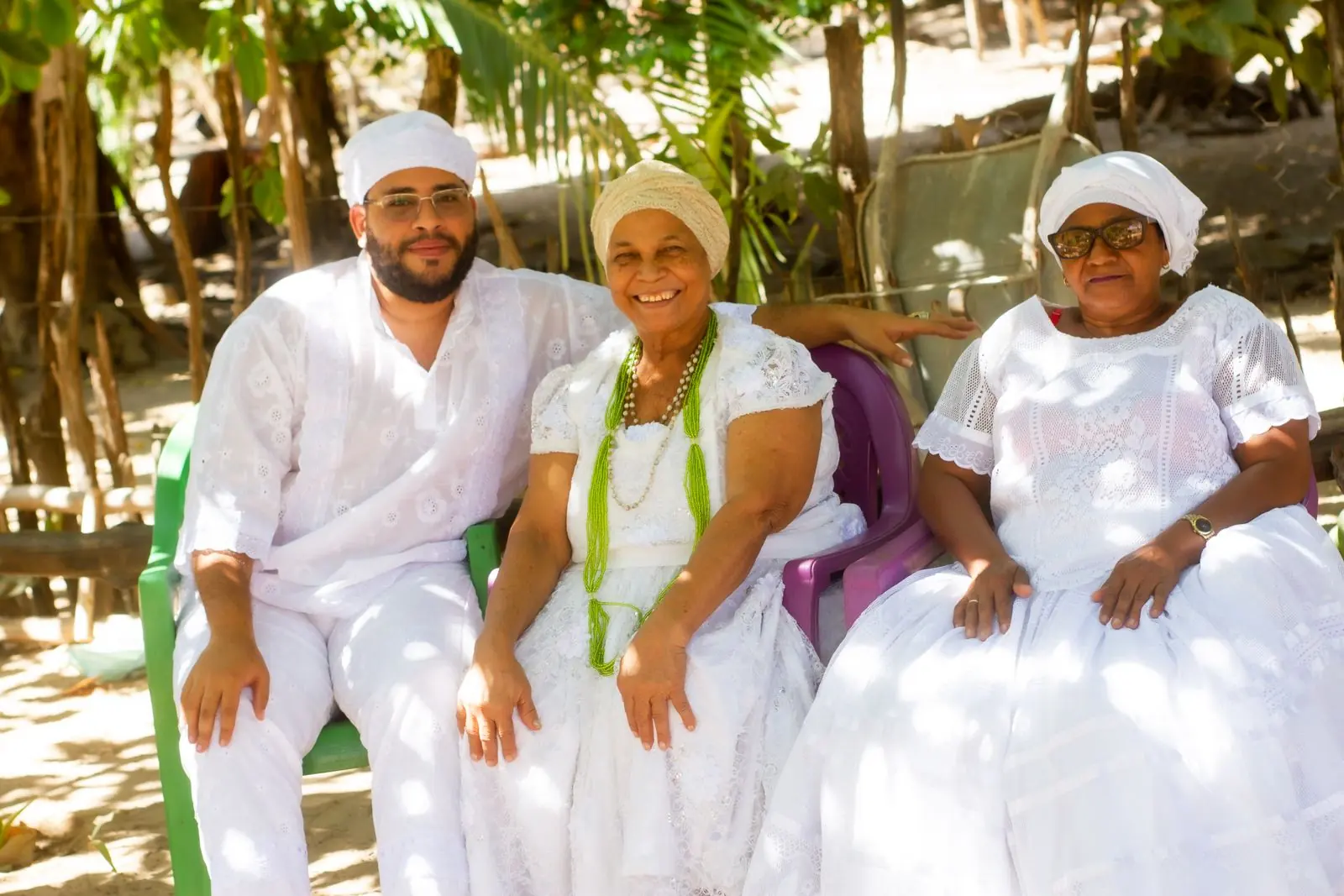 Três integrantes da corrente vestidos de branco, sorrindo, sentados no quintal do terreiro